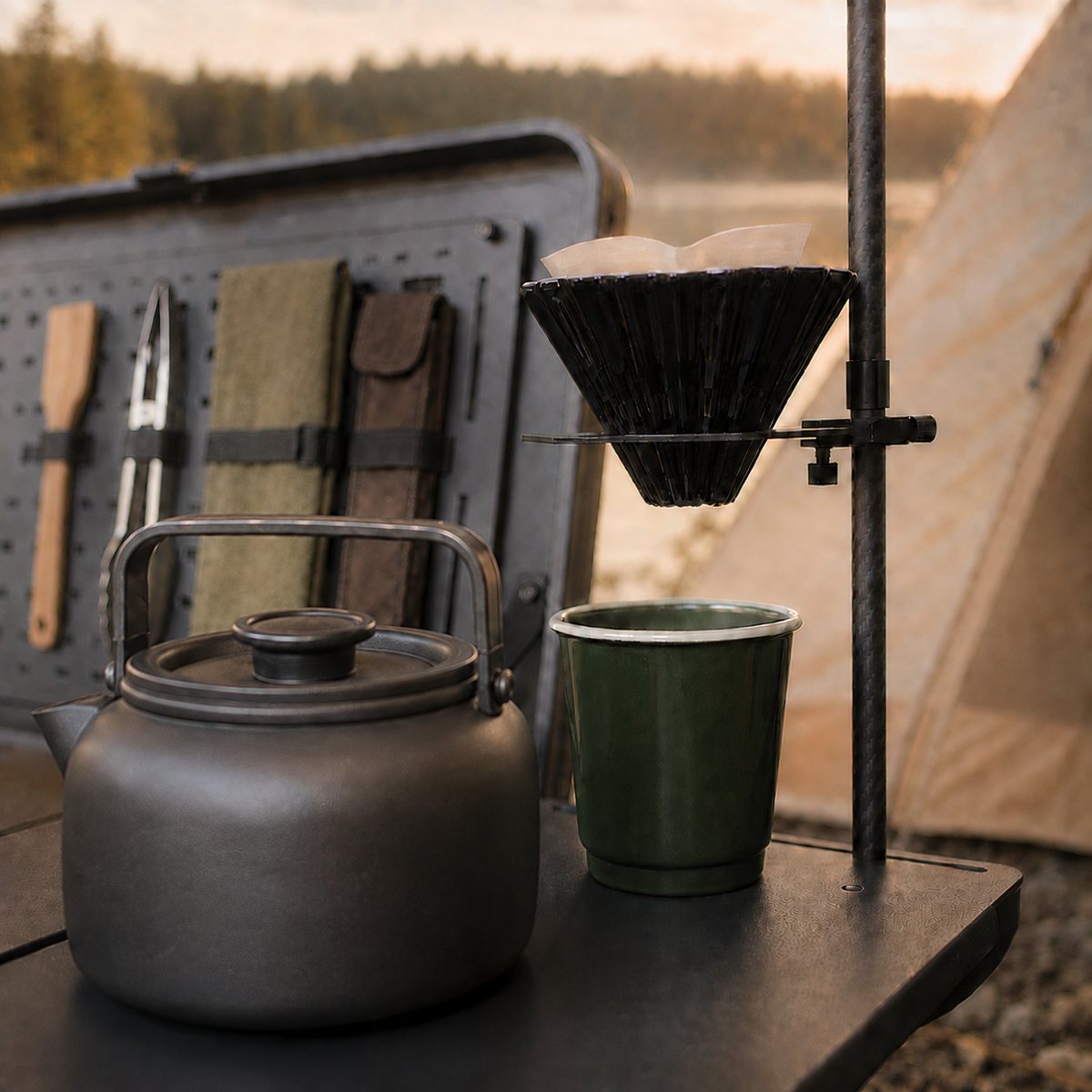 Kaffeehalter der Rauholm Campküche 50 mit Filteraufsatz, daneben Wasserkessel und Emaillebecher auf der Arbeitsfläche, im Hintergrund der Deckel mit Kochwerkzeug